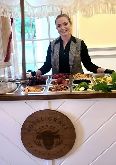 Smiling woman behind a grazing table with fruits and snacks at Royal Graze Charcuterie.