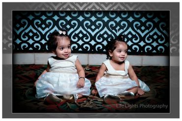 Two adorable toddlers in white dresses sitting on a patterned carpet.