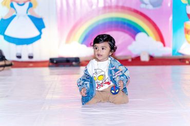 A toddler in colorful clothes sitting on a shiny floor with a rainbow backdrop.