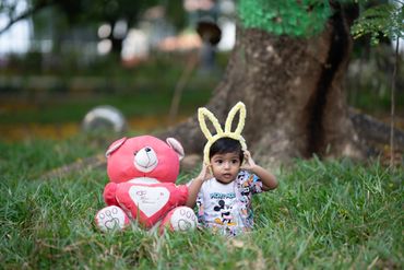A child with bunny ears sitting on grass next to a pink teddy bear.