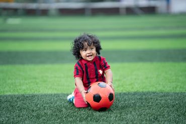 A happy toddler with curly hair plays with a red and black soccer ball on a green field.