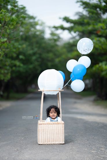 A baby sitting in a wicker basket with blue and white balloons.