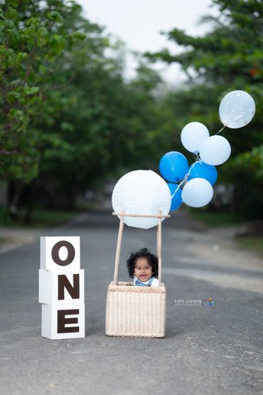 Happy baby in a basket with balloons beside blocks spelling ONE.