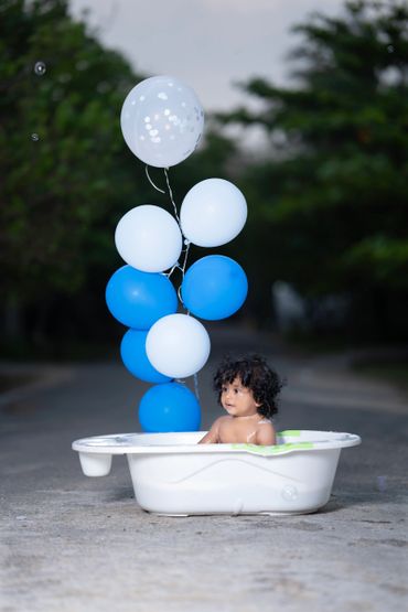 A baby sitting in a white tub with blue and white balloons outdoors.
