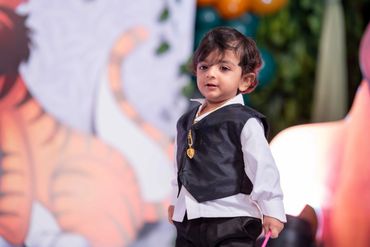 A young boy dressed in a formal black vest and white shirt at a colorful event.