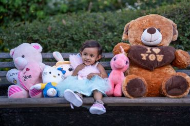 A little girl sits happily on a bench surrounded by colorful stuffed animals.
