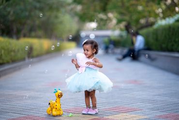 A little girl in a blue dress playing with bubbles and a yellow toy giraffe.