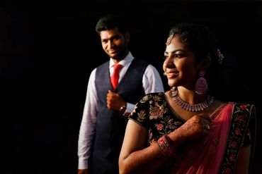 A smiling woman in traditional attire with a man in formal wear in the background.