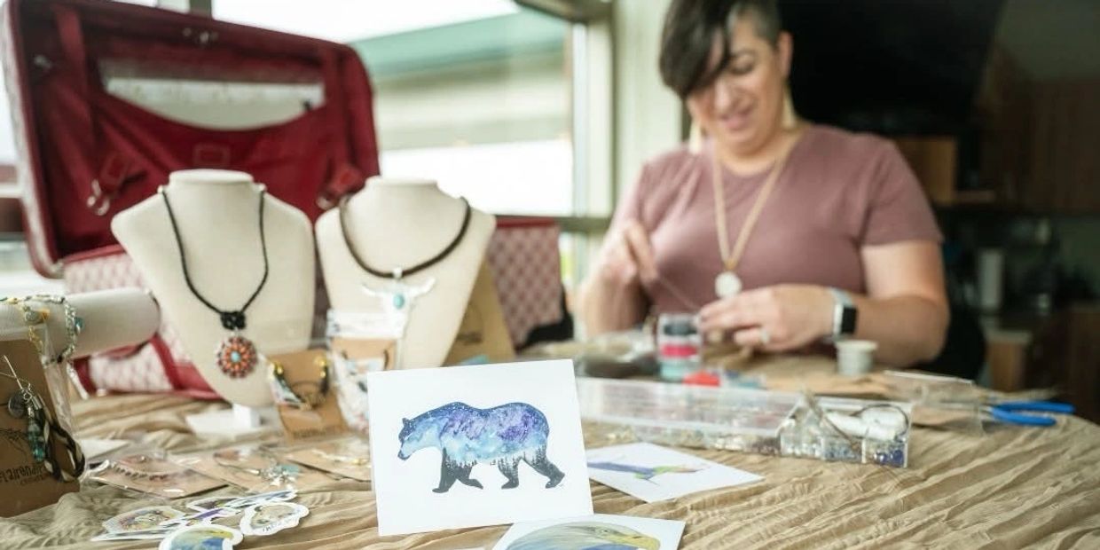 Woman crafting jewelry with a display of necklaces and art cards on a table.