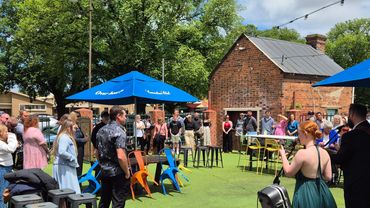 Outdoor gathering with people standing and chatting under blue umbrellas on a sunny day.