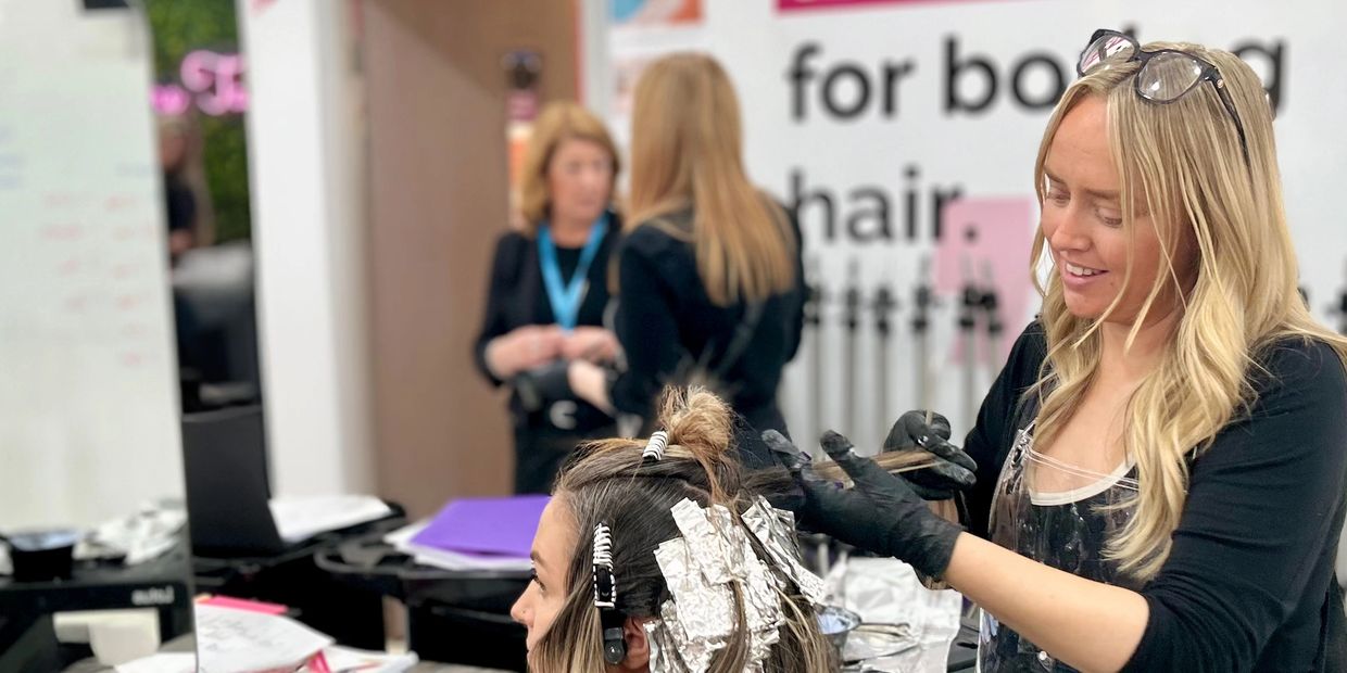 A female hairdresser takes out hair strands from client to apply foil. Educators talk in background.