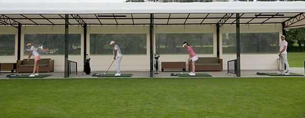 Four people practicing golf swings at a driving range with a covered shelter.