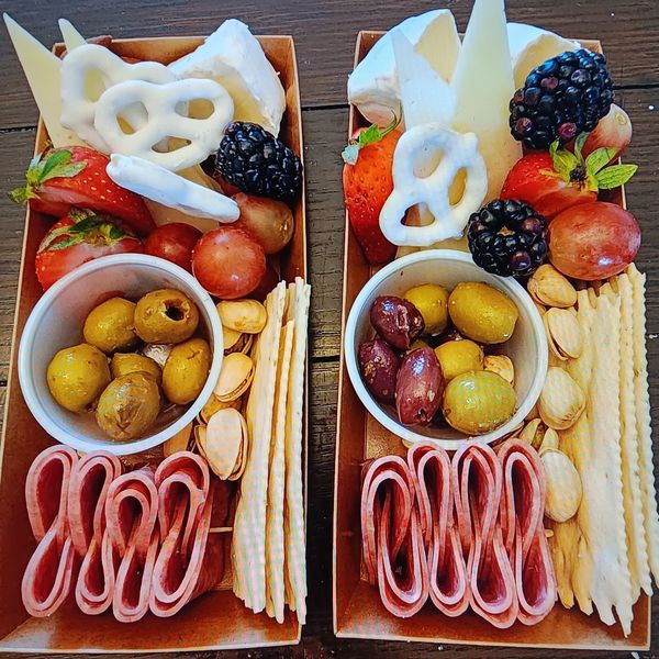 Two charcuterie boxes with fruits, cheese, crackers, olives, and nuts.
