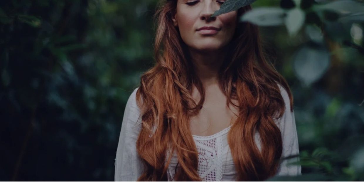 Woman with long red hair in a natural, leafy setting, eyes closed in calm.