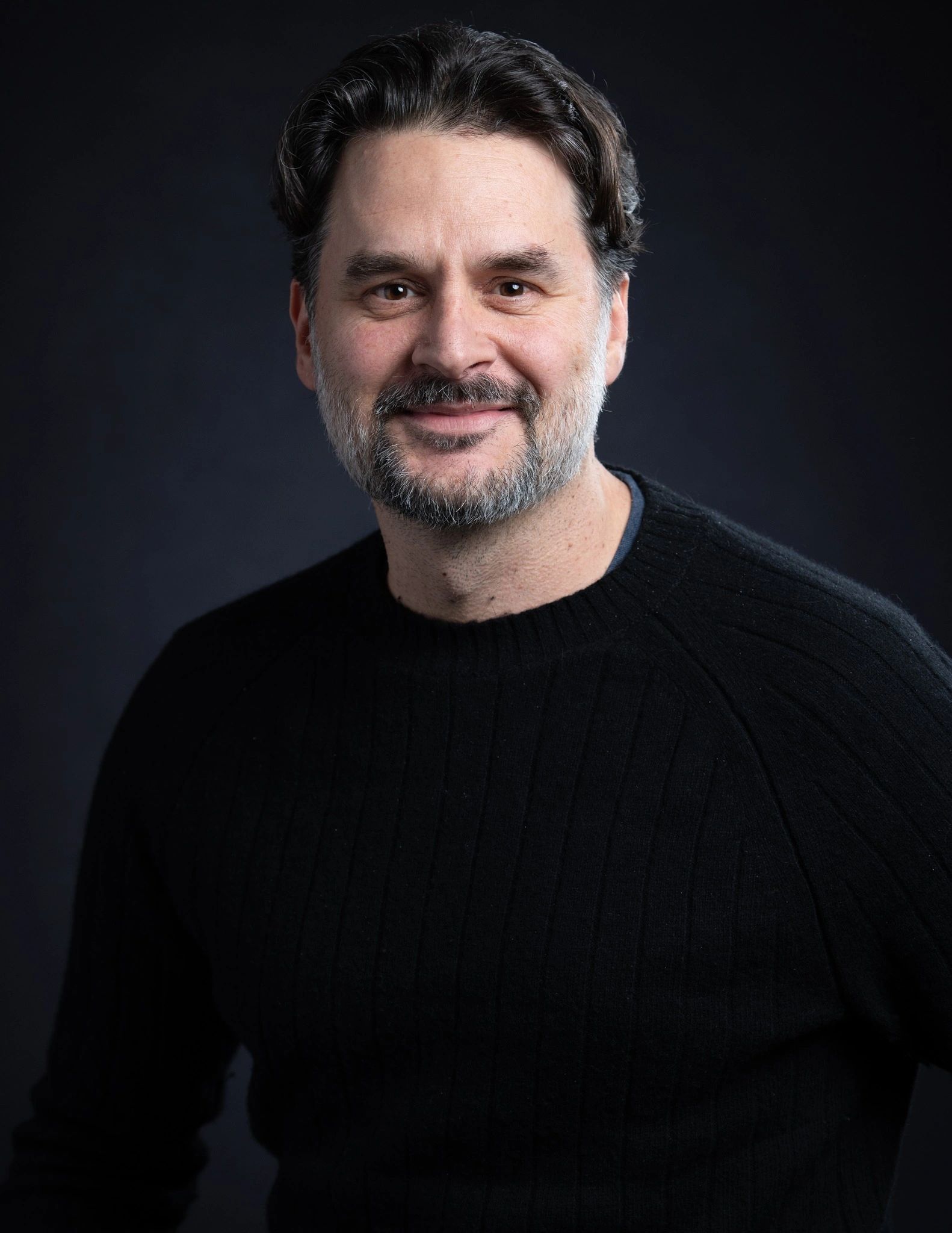 Smiling man with dark hair and beard wearing a black sweater against a dark background.