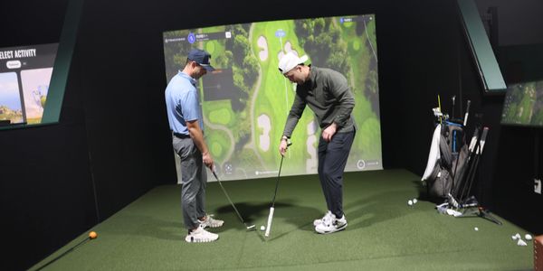 Two men practicing golf indoors with a simulator and putting green.