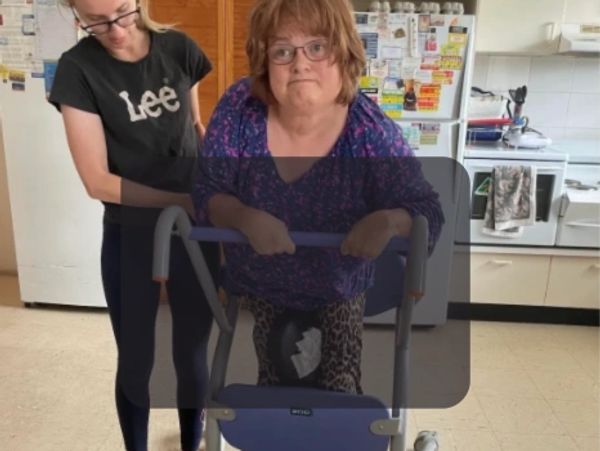 A woman with a walker is assisted by another woman in a kitchen setting.