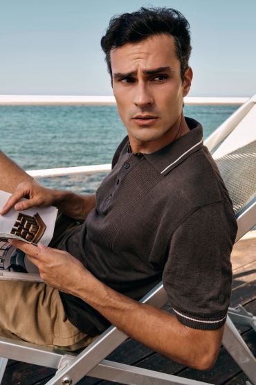 Man in a dark polo shirt sitting on a deck chair by the sea, holding a magazine.
