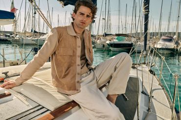 Man in beige jacket sitting on a sailboat docked at a marina.