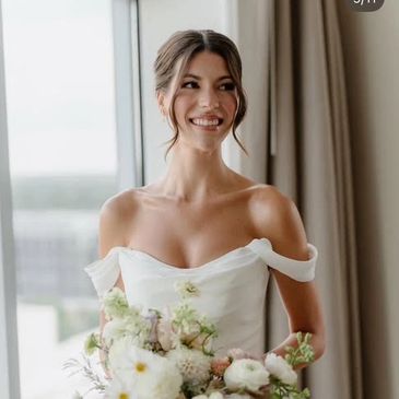 Bride in a white off-shoulder dress holding a bouquet, smiling.