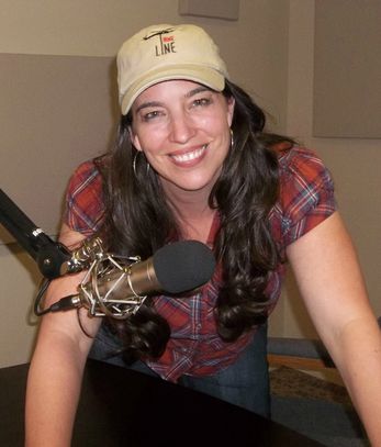 Smiling woman wearing a cap and plaid shirt speaking into a microphone.