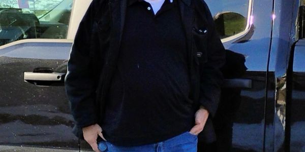 Person in black jacket and jeans standing by a black vehicle.