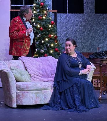 Two people in elegant attire near a decorated Christmas tree on stage.