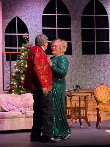 Couple in festive attire dancing on stage with Christmas tree background.