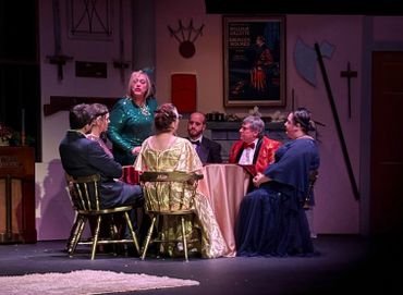 Actors in formal attire perform a dramatic scene around a table on stage.