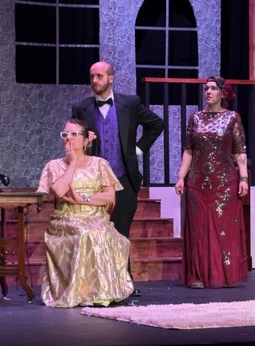 Three actors in formal vintage attire performing on stage with a stone wall backdrop.