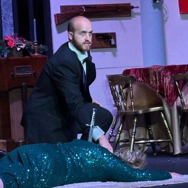 A man kneels beside a woman lying on the floor with a sword through her back on stage.