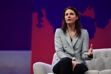 Woman in a checkered blazer speaking on stage with a purple and red backdrop.