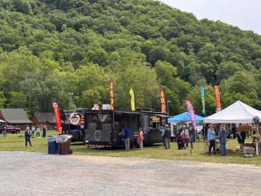 People enjoy an outdoor food truck and artisan market surrounded by trees.