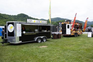 Two food trucks, one selling lemonade, set on grassy field with mountains in background.