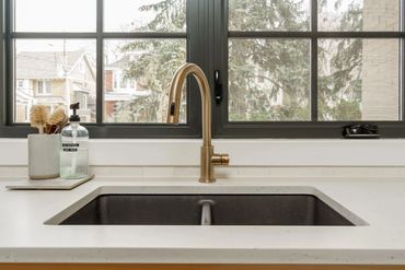 Gold kitchen faucet and sink with granite countertop in front of a window with dark frames