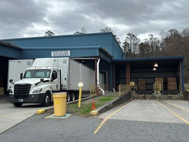 A white semi-truck parked at a loading dock of a blue warehouse on a cloudy day.