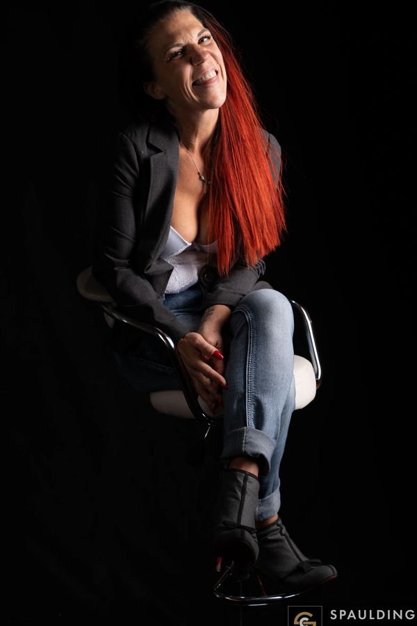 Smiling woman with vibrant red hair sitting on a stool against a black background.
