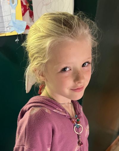 Young girl with blonde hair smiling softly, wearing a purple hoodie and colorful necklace.