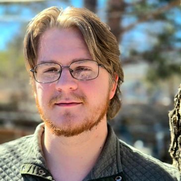Young man with glasses and blonde hair smiling outdoors.