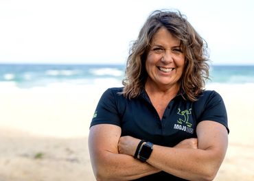 Smiling woman with curly hair in black Mojo 365 shirt at the beach.