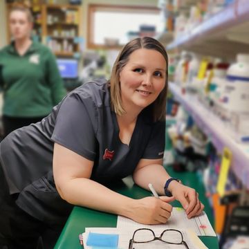 Woman in grey scrubs writing on paper in pharmacy.