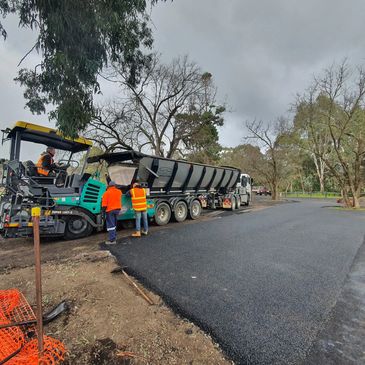 Workers paving a new asphalt road with heavy machinery in a wooded area.