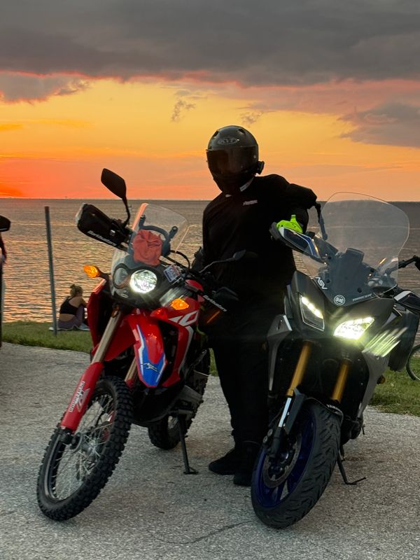 Motorcyclist posing with two bikes at sunset by the water.