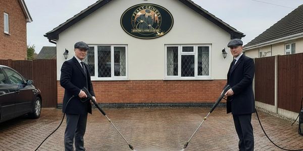 Two men in Peaky Blinders attire power-washing a driveway.