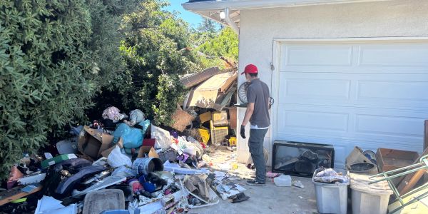 A man in a red cap stands near a large pile of trash outside a house.