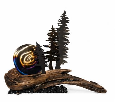 Artistic sculpture combining metal trees, colorful disc, and driftwood on black stones.