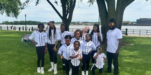 A group of girls and a man posing in matching sports-style white shirts by a river.