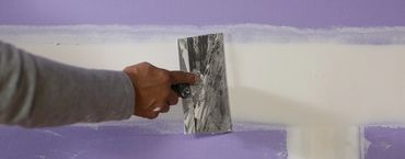 Hand using a putty knife to smooth white joint compound on a purple drywall.