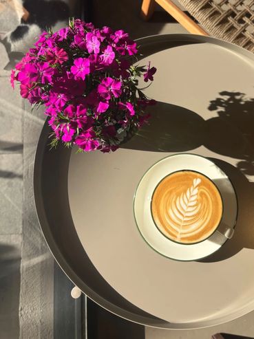 A cup of cappuccino with latte art next to vibrant purple flowers on a round table.