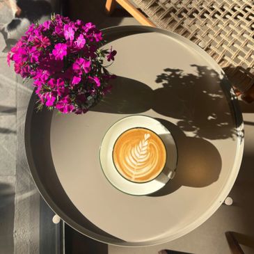 A cup of latte with leaf latte art and purple flowers on a round table.